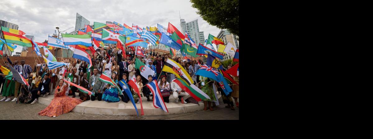 A large group of One Young World delegates are sitting and standing waving their home nation flags outside a conference venue,