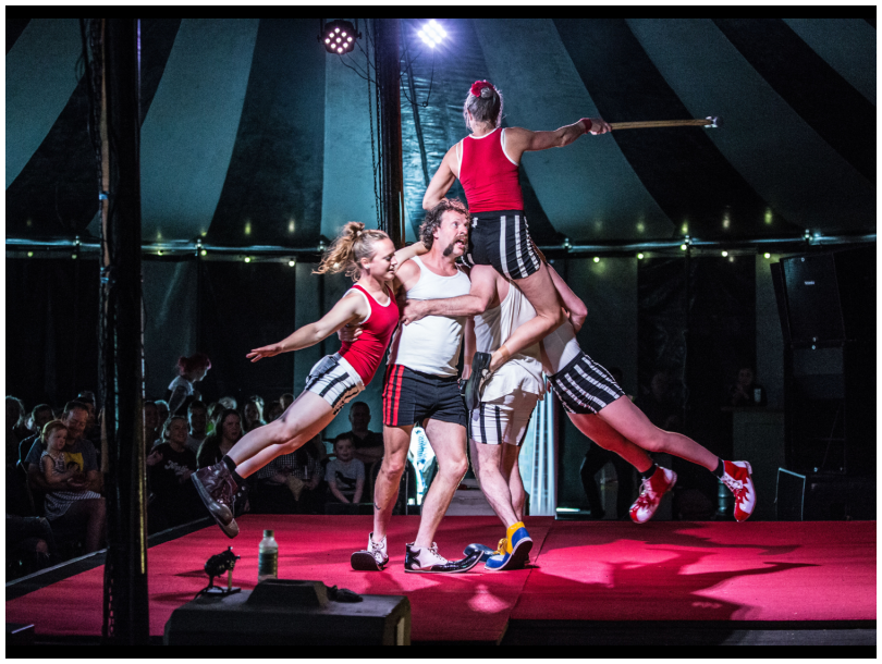 Five circus performers performing feats of acrobatic magic with three performers hanging off two male performers as they spin around on a red stage. 
