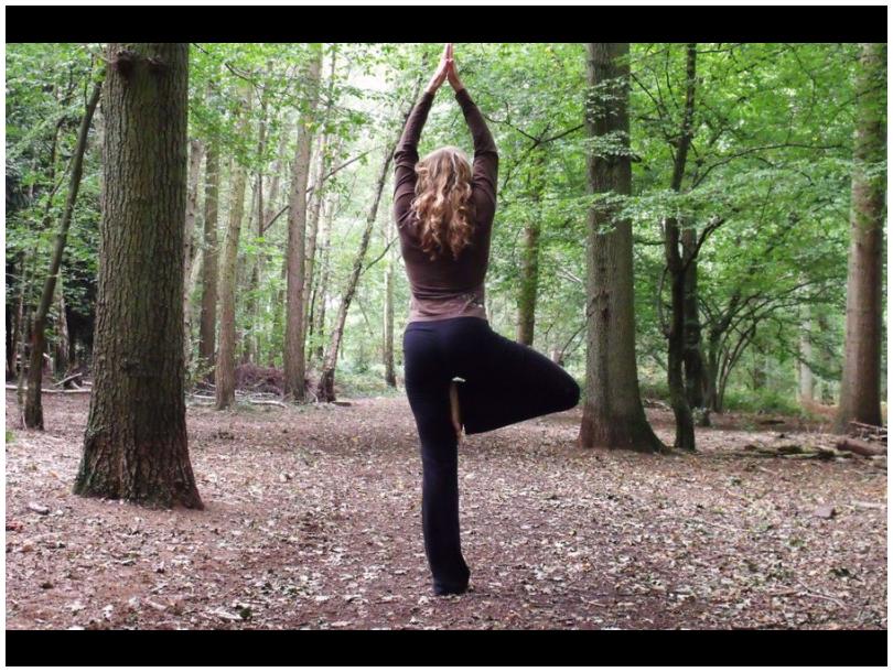 A lady doing yoga in the woods, on one leg doing tree pose.