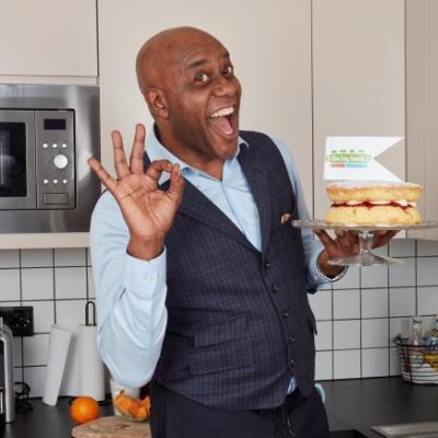 ainsley harriot with big lunch flag and cake looking delighted and holding fingers up  to make ok sign