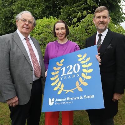 Three people hold up a sign celebrating 120 years of James Brown & Sons